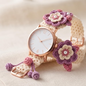 Può includere: Un orologio da polso con cassa color oro rosa e quadrante bianco con sottili indicatori. Il cinturino è realizzato in tessuto all'uncinetto color crema, decorato con fiori all'uncinetto viola e crema, piccole perline bianche e sfere viola.