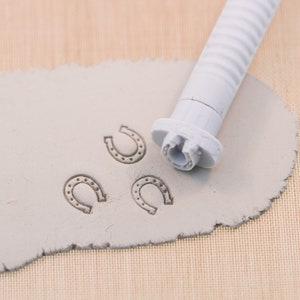 May include: A gray horseshoe stamp tool next to three stamped horseshoe shapes on a gray clay surface. The stamp tool has a ridged handle and a horseshoe-shaped stamping head. The stamped horseshoes have a dotted pattern.