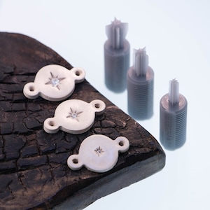 Może przedstawiać: Trzy białe ceramiczne zawieszki do biżuterii z wzorem gwiazdy i małym, przezroczystym kryształem w środku. Zawieszki leżą na ciemnym drewnianym podłożu. W tle znajdują się trzy białe plastikowe formy.