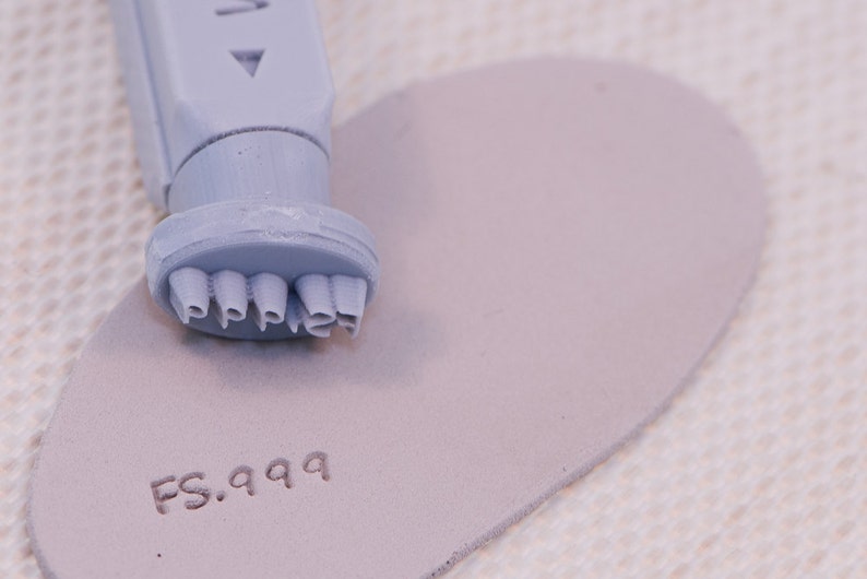 May include: A gray rubber stamp with the word "PIPPI" embossed on the bottom. The stamp is on a light gray piece of leather with the text "FS.999" stamped on it.