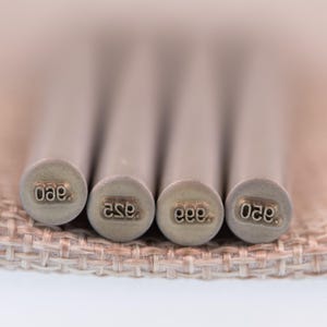 May include: Four cylindrical metal stamping tools arranged side by side. Each tool features a circular end engraved with specific numerical markings, including 950, 925, 999, and 850, designed for marking precious metal jewellery pieces.