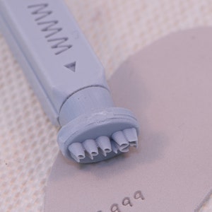 May include: A gray plastic stamp with the word "Pepper" embossed on the bottom. The stamp is being used to stamp the words "FS.999" onto a piece of gray fabric.