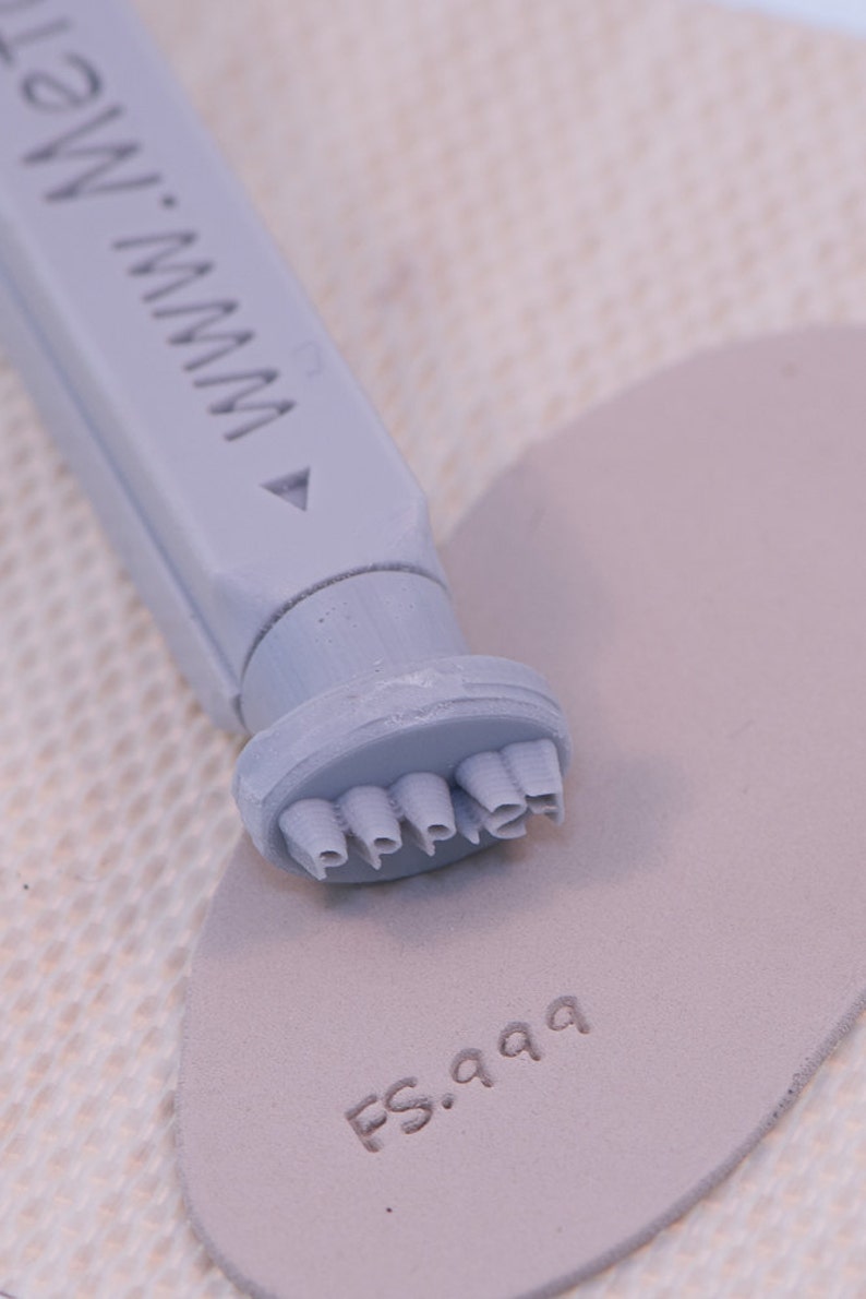 May include: A gray plastic stamp with the word "Pepper" embossed on the bottom. The stamp is being used to stamp the words "FS.999" onto a piece of gray fabric.
