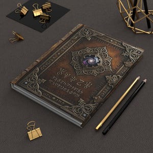 Puede incluir: Un diario de tapa dura rectangular con una cubierta marrón de aspecto desgastado, con bordes de filigrana plateada, un emblema circular central con temática de galaxia y filas de texto blanco con estilo de runas. Dos lápices y clips dorados.