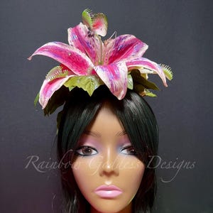 Op de afbeelding: Een hoofddeksel met een grote, levendige roze en witte lelie omringd door groene bladeren en kleine Venusvliegenvanger-accenten. Het bloemstuk zit op een etalagepophoofd met lang zwart haar. De tekst Rainbow Goddess Designs is zichtbaar.