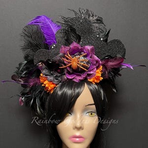 May include: A Halloween-themed headpiece with black feathers, purple accents, and orange and purple flowers. A glittery black bat and spider are also featured. The text "Rainbow Goddess Designs" is visible.