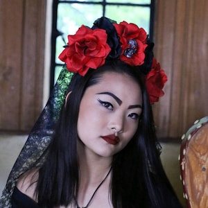 Op de afbeelding: Een persoon draagt een zwarte off-the-shoulder top met de tekst "DEARLY DEPARTED" en een bloemenkrans van rode en zwarte rozen. Een zwarte kanten sluier is over haar hoofd gedrapeerd en een kogelhanger hangt aan een ketting.