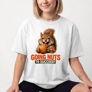Puede incluir: Una camiseta blanca de manga corta con un gráfico de una ardilla marrón sosteniendo una nuez. Debajo de la ardilla, el texto "GOING NUTS TO SUCCEED!" aparece en letras negritas de color naranja y negro. La camiseta tiene cuello redondo y corte holgado.