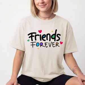 May include: A beige short-sleeved t-shirt featuring the black text "Friends FOREVER" printed on the front. The design includes small, colourful hearts in red, yellow, green, and blue scattered around the lettering, with the letter O filled in blue.