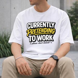 May include: A white short-sleeved t-shirt featuring the bold black and yellow text "CURRENTLY PRETENDING TO WORK" followed by the smaller black text "please don't disturb" with decorative wavy lines on either side of the bottom phrase.