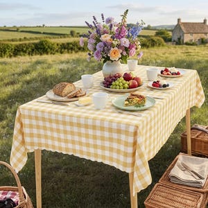May include: A rectangular table covered with a yellow and white gingham tablecloth, set with a ceramic vase of colorful flowers, plates of sliced bread, fresh fruit, a slice of quiche, butter, and white cups, positioned outdoors in a grassy field.