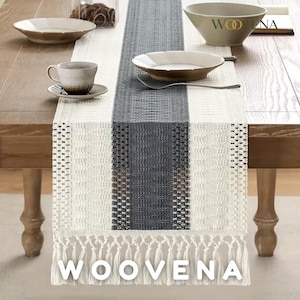 May include: A rectangular table runner featuring a central dark gray woven panel flanked by cream-colored sections with intricate textured patterns and open-weave details. The runner ends in long, knotted cream fringe. The text WOOVENA is visible.