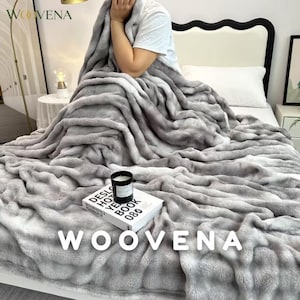 Puede incluir: Una manta de textura suave con degradado gris y blanco sobre una cama. Una vela negra en un frasco de vidrio descansa sobre un libro blanco titulado DESLO HOTEL YEAR BOOK 080, colocado sobre la superficie de la manta de piel sintética.