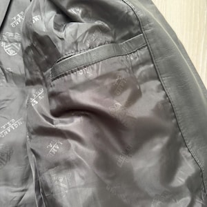 May include: Close-up of the interior of a black leather jacket. The lining is a dark gray with a repeating pattern and the text "ENGLAND U.S.A.". A pocket is visible on the left side.