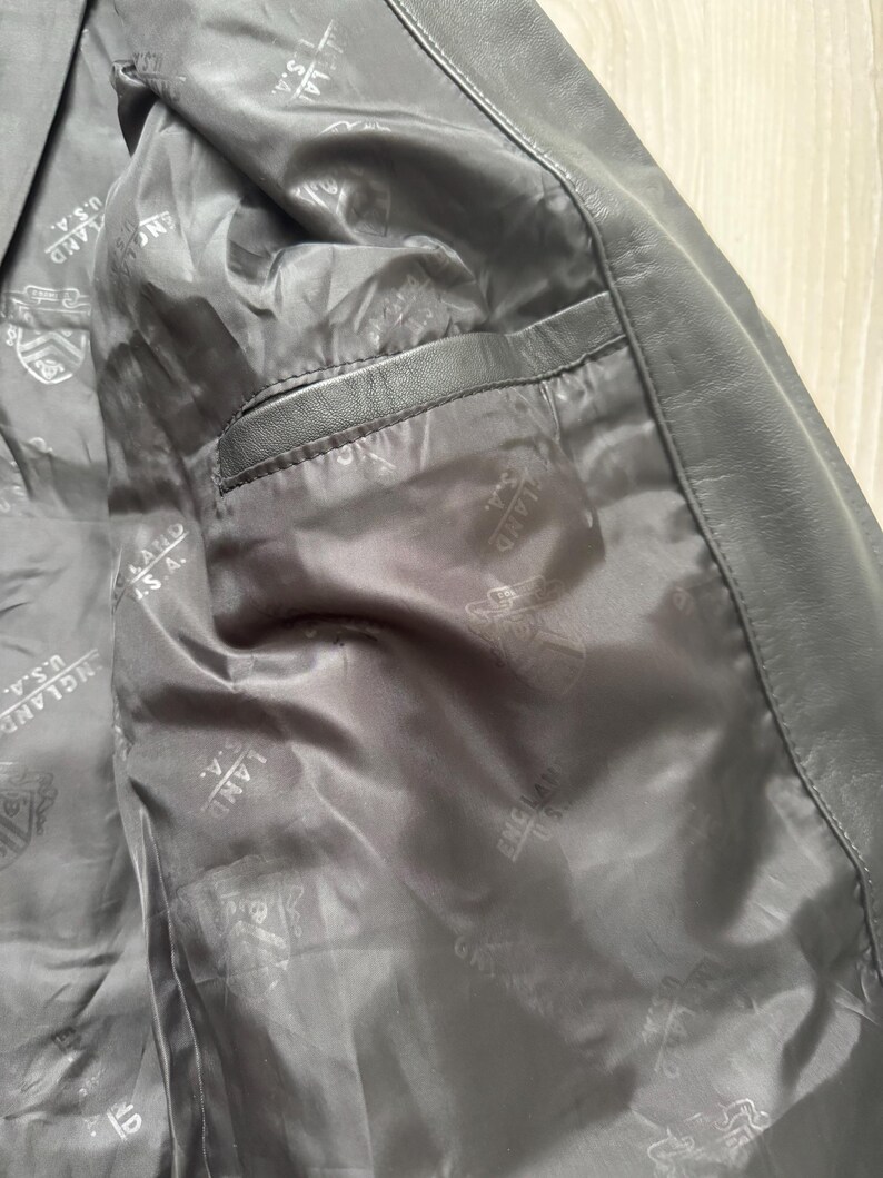 May include: Close-up of the interior of a black leather jacket. The lining is a dark gray with a repeating pattern and the text "ENGLAND U.S.A.". A pocket is visible on the left side.