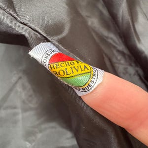May include: Close-up of a tag with the Bolivian flag colors: red, yellow, and green. The tag reads "HECHO EN BOLIVIA" in a circular design. The tag is attached to a dark gray fabric.