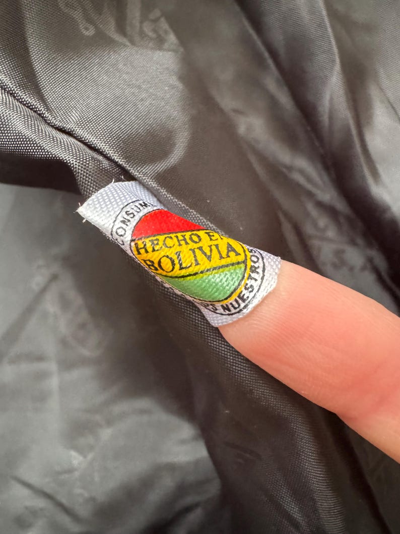 May include: Close-up of a tag with the Bolivian flag colors: red, yellow, and green. The tag reads "HECHO EN BOLIVIA" in a circular design. The tag is attached to a dark gray fabric.