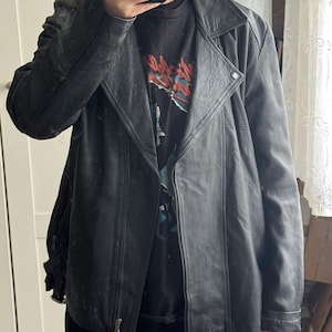 May include: A black leather jacket with an asymmetrical zipper closure and a notched collar. The jacket has a classic biker style with a slightly worn appearance. The model is wearing a black graphic t-shirt underneath.