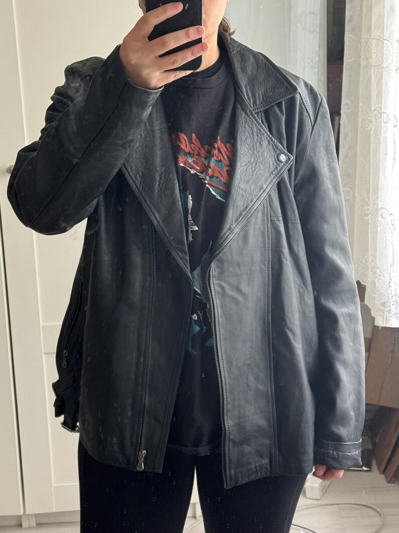 May include: A black leather jacket with an asymmetrical zipper closure and a notched collar. The jacket has a classic biker style with a slightly worn appearance. The model is wearing a black graphic t-shirt underneath.