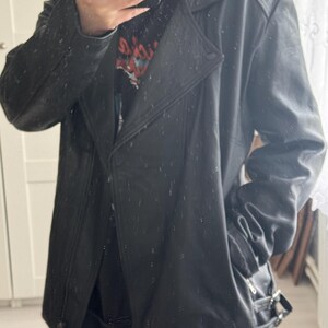 May include: Black leather jacket with a notched collar, asymmetrical zipper closure, and a belt detail at the waist. The jacket has a classic biker style with a slightly oversized fit. The jacket has a button closure and a zipper pocket.