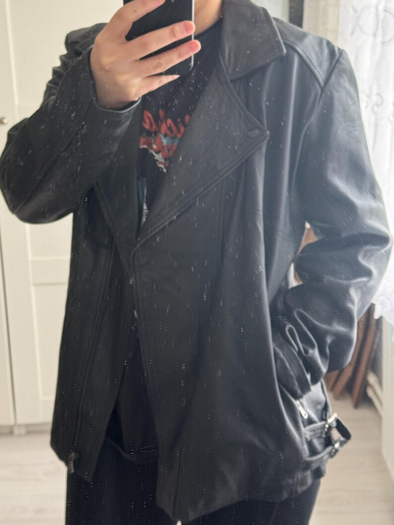 May include: Black leather jacket with a notched collar, asymmetrical zipper closure, and a belt detail at the waist. The jacket has a classic biker style with a slightly oversized fit. The jacket has a button closure and a zipper pocket.