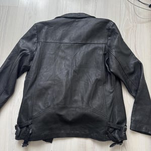 May include: Black leather jacket with a classic design. Features include a notched collar, shoulder epaulets, and adjustable side buckles. The jacket has a smooth texture and a tailored fit, suitable for various styles.