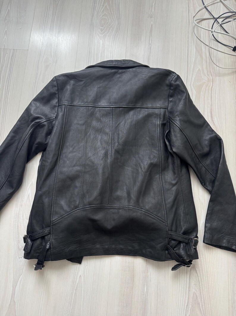 May include: Black leather jacket with a classic design. Features include a notched collar, shoulder epaulets, and adjustable side buckles. The jacket has a smooth texture and a tailored fit, suitable for various styles.