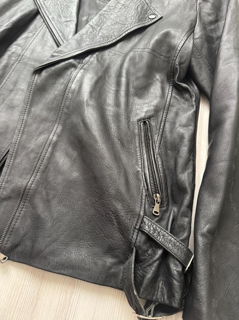 May include: Black leather jacket with an asymmetrical zipper closure, a notched lapel, and a zippered chest pocket. The jacket has a belt detail at the waist and a classic biker style.