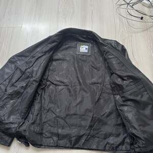 May include: Black leather jacket with an open front, revealing a dark interior lining. The jacket has a classic design with a collar and a small label on the inside. The leather appears worn, suggesting a vintage style.