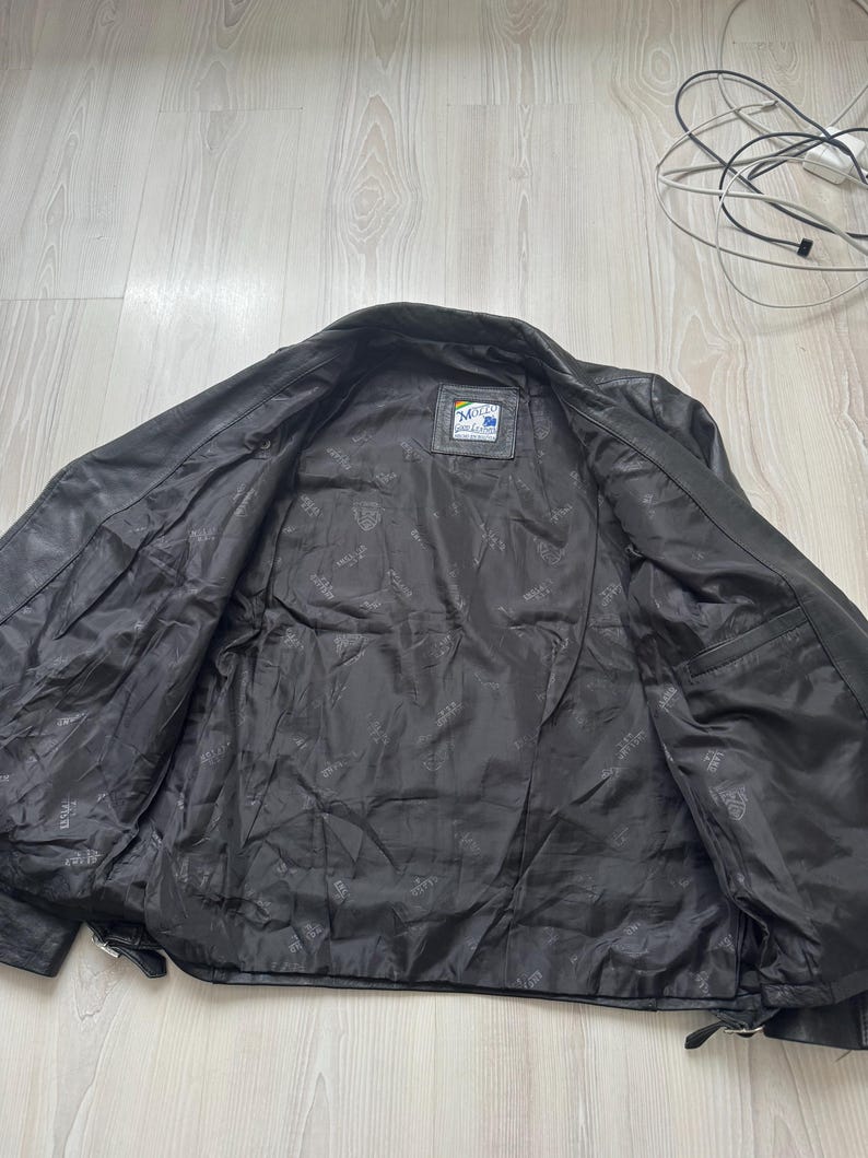 May include: Black leather jacket with an open front, revealing a dark interior lining. The jacket has a classic design with a collar and a small label on the inside. The leather appears worn, suggesting a vintage style.