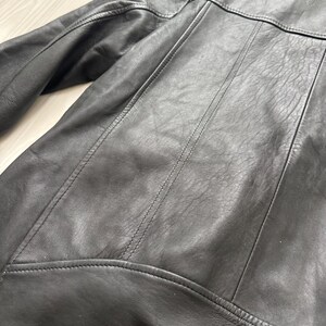 May include: Close-up of a black leather jacket. The jacket features detailed stitching along the seams, a collar, and shoulder panels. The leather has a slightly textured appearance.