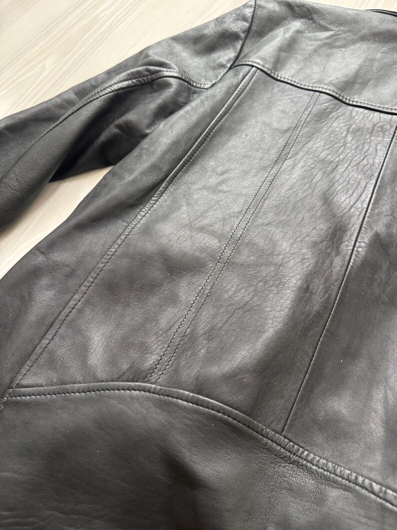 May include: Close-up of a black leather jacket. The jacket features detailed stitching along the seams, a collar, and shoulder panels. The leather has a slightly textured appearance.