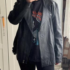 May include: Black leather jacket with a notched collar, asymmetrical zipper, and a snap detail at the shoulder. The jacket is worn over a black graphic t-shirt and black leggings. The jacket has a classic biker style.