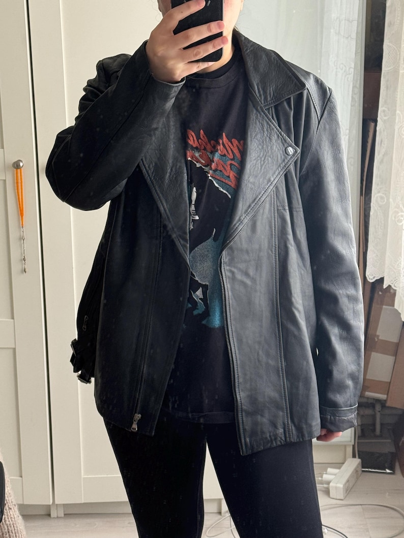 May include: Black leather jacket with a notched collar, asymmetrical zipper, and a snap detail at the shoulder. The jacket is worn over a black graphic t-shirt and black leggings. The jacket has a classic biker style.
