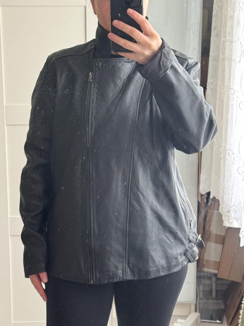 May include: Black leather jacket with an asymmetrical zipper closure and a high collar. The jacket has a fitted silhouette, long sleeves, and a buckle detail at the waist. The jacket appears to be made of a smooth, possibly genuine leather material.