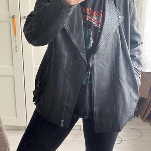May include: A black leather jacket with a zipper closure and a notched collar. The jacket has a slightly distressed appearance and is worn over a graphic t-shirt and black leggings. The jacket has a classic biker style.