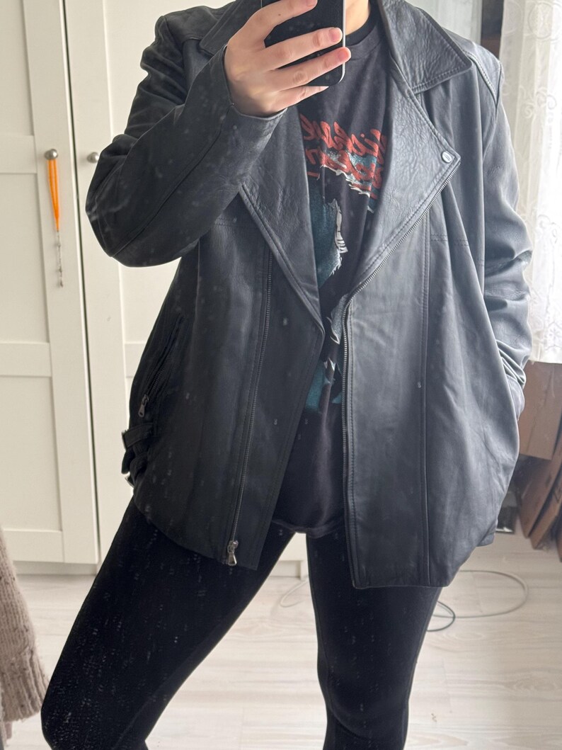 May include: A black leather jacket with a zipper closure and a notched collar. The jacket has a slightly distressed appearance and is worn over a graphic t-shirt and black leggings. The jacket has a classic biker style.