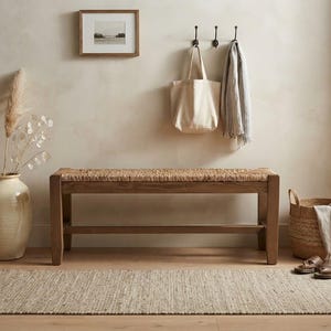 May include: A rectangular wooden bench with a woven rush seat, positioned in a room with a large cream-colored ceramic vase containing dried pampas grass, a woven basket with a light-colored cloth, and a textured beige area rug on the floor.