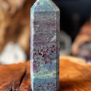 Peut inclure: Une tour en cristal hexagonale présentant un mélange tacheté d'inclusions minérales magenta profond, vert forêt et bleu ardoise. La pierre possède une surface polie et lisse avec un sommet pointu et une base plate, révélant des nuances naturelles.