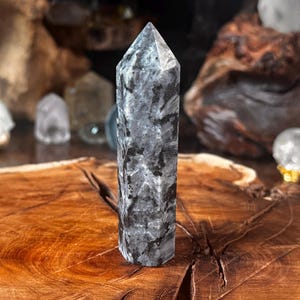 May include: A polished, hexagonal crystal tower featuring a mottled pattern of dark charcoal grey, black, and translucent light grey mineral inclusions. The stone has a smooth, faceted surface with a pointed top and a flat base, displaying natural variations.