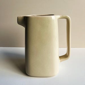 Puede incluir: Una jarra de cerámica de color crema con un acabado liso y brillante. El recipiente presenta un cuerpo alto y rectangular con esquinas redondeadas, un pico vertedor ancho en el lado izquierdo y una gran asa angular en el lado derecho.