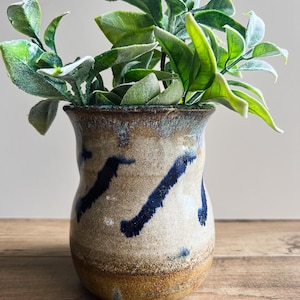 May include: A ceramic vase with a speckled beige and tan glaze featuring diagonal dark blue brushstroke patterns. The vase has a flared rim and a rounded body, containing a green artificial plant with multiple leaves extending upwards.