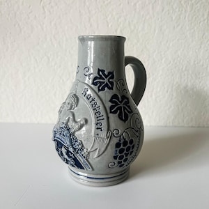 May include: A gray ceramic pitcher featuring a blue relief design of grapevines and a figure holding a barrel. A banner across the front displays the text Ratskeller in blue lettering. The pitcher has a curved handle and a smooth, glazed finish.