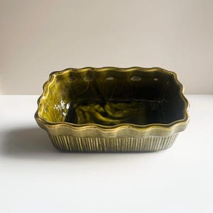 May include: A rectangular ceramic baking dish with a glossy olive green glaze. The exterior features vertical ribbed detailing, while the upper rim has a scalloped, wavy edge. The interior is smooth and deep, reflecting light on its dark green surface.