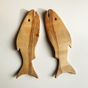 May include: Two fish-shaped wooden boards with a natural light brown wood grain pattern. Each board features a cutout near the top to represent an eye and a smooth, contoured silhouette resembling a fish with a defined tail and fin shape.