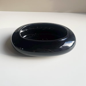 May include: A glossy black ceramic bowl with an oval shape and a smooth, reflective surface. The bowl features a wide, rounded rim and a deep, hollow center, presenting a clean, solid color design with no additional patterns or decorative elements.