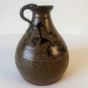 May include: A brown ceramic pitcher with a speckled glaze finish and a single handle. The body of the pitcher features a hand-painted black vine design with swirling tendrils and leaves that wrap around the front of the rounded vessel.
