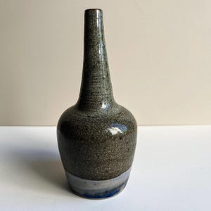 May include: A ceramic bud vase with a tall, slender neck and a rounded body. The upper portion features a speckled, dark olive-green glaze, while the base transitions into a smooth, light grey band with a subtle hint of blue at the very bottom edge.