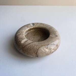 May include: A circular ashtray crafted from polished beige and brown marbled stone. The object features a smooth, rounded rim with three distinct notches spaced evenly around the perimeter, leading into a deep, concave center basin.