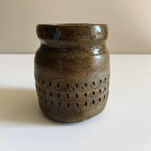 May include: A small, cylindrical ceramic vessel with a glossy, mottled brown glaze. The exterior features a textured band of evenly spaced, vertical indentations encircling the lower portion of the body, creating a subtle, repeating pattern.